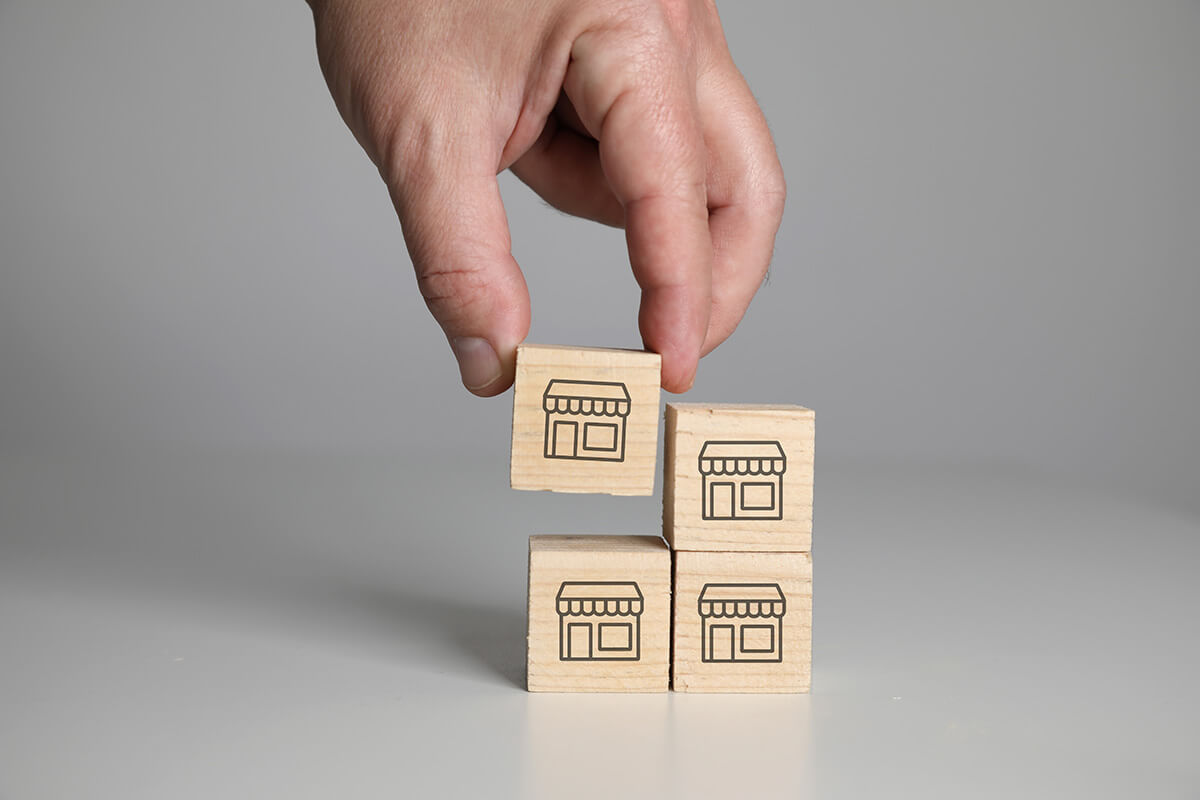 A hand places a wooden block depicting a shop onto a stack of three similar blocks on a white surface. This visual represents the growth and expansion possible when you franchise your business with professional guidance from franchise consulting firms.