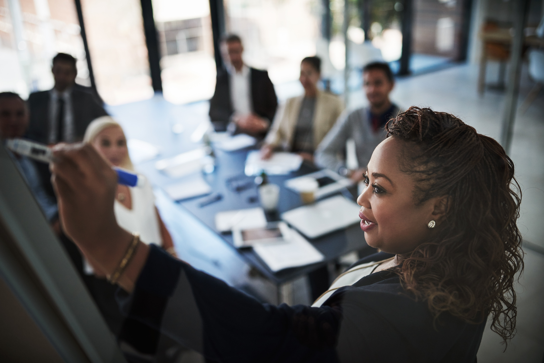 doing a business presentation|A woman writes on a transparent board while six colleagues sit at a table watching attentively during a meeting in a modern office space