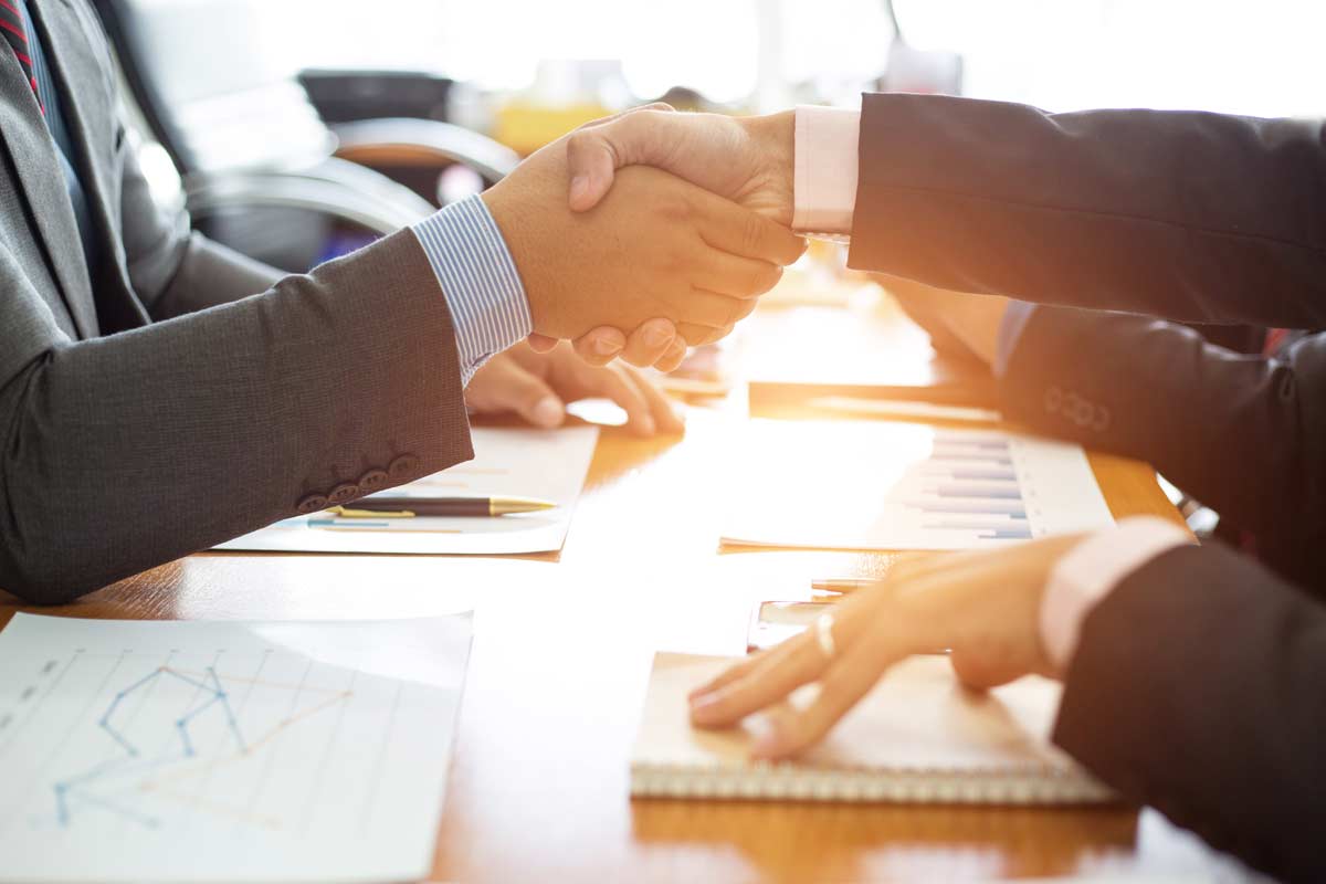 Two people in suits shake hands over a table with documents